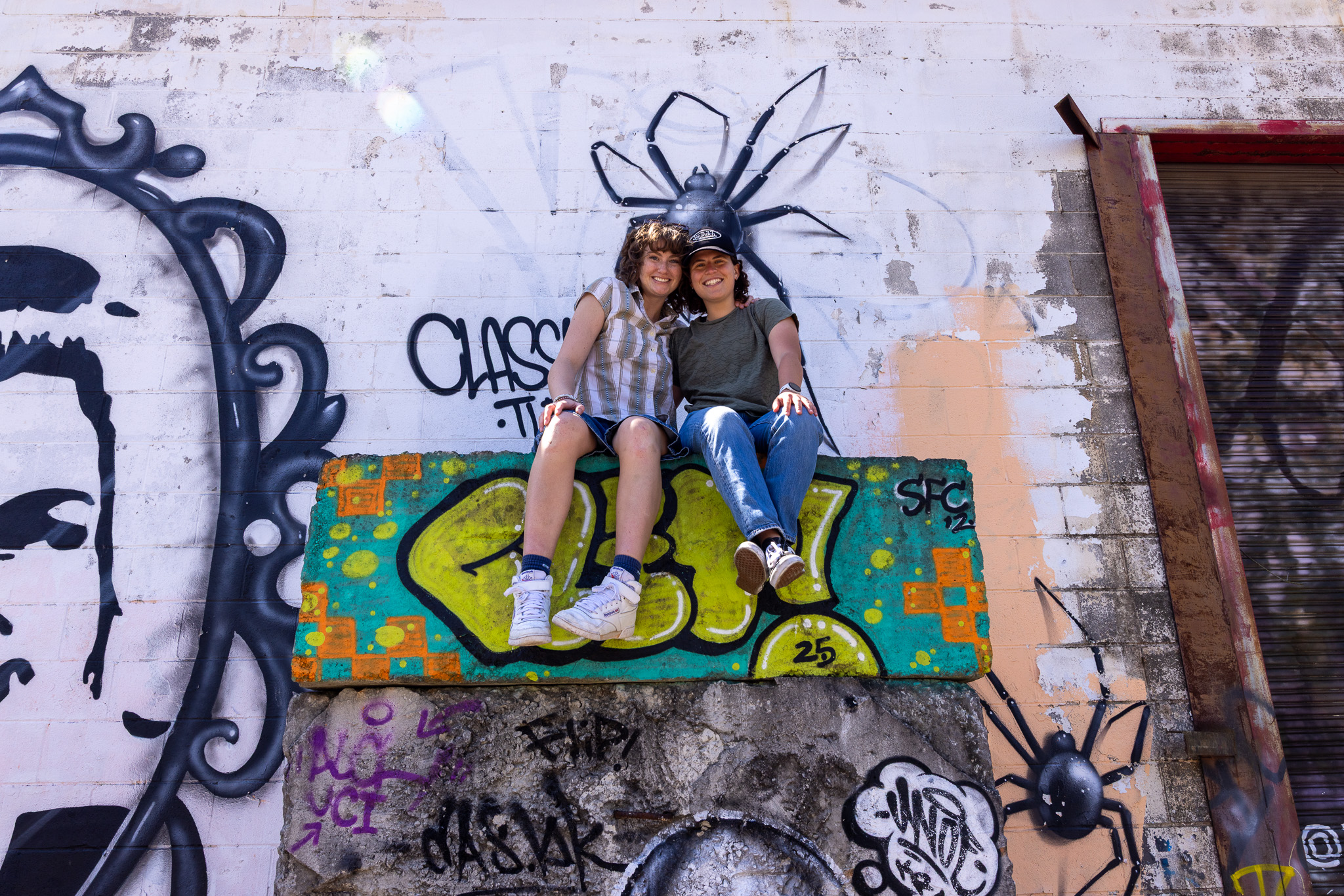 Two people sitting up high on a warehouse structure in front of a brightly painted mural
